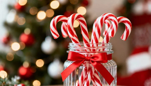Kırmızı ve beyaz çizgili baston şekerlerle dolu cam bir kavanozun yakın plan fotoğrafı, kırmızı kurdeleyle bağlanmış, bulanık bir Noel ağacı arka planına yaslanmış, ve sıcak bokeh ışıkları, rahat ve neşeli bir tatil sahnesi yaratıyor..