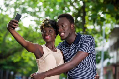 Genç bir çift parkta durup gülümserken cep telefonuna bakıyor..