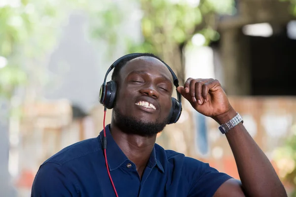 年轻人在白天坐在外面的椅子上 一边用耳机听音乐 微笑的人闭着眼睛听音乐 — 图库照片