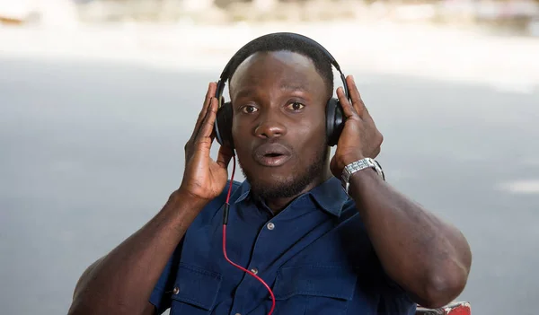 Młody Człowiek Siedzący Zewnątrz Słuchający Muzyki Pomocą Słuchawki Patrzący Kamerę — Zdjęcie stockowe