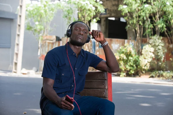 young man listens to music with headphones while sitting on a chair outside in the day.