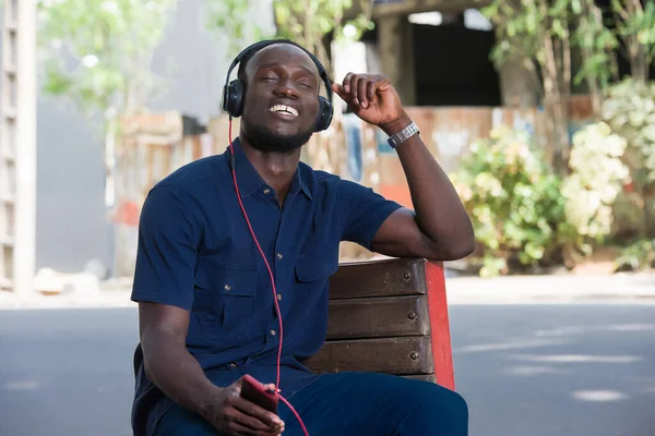 young man listens to music with headphones while sitting on a chair outside in the day. smiling man listens to music with his eyes closed.