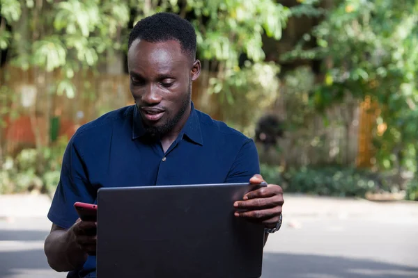 Молодий Чоловік Сидить Відкритому Повітрі Ноутбуком Дивлячись Мобільний Телефон Посміхаючись — стокове фото