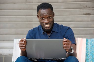 Genç adam alışveriş torbaları ve dizüstü bilgisayarla yerde oturmuş kameraya bakıp gülüyor..