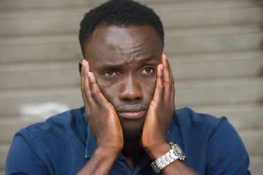 Mavi tişörtlü genç adam dışarıda oturuyor ve ellerin yanaklarda olduğunu düşünüyor..