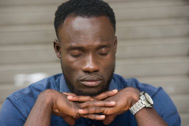 Açık havada oturan mavi gömlekli genç adam kapalı gözleri düşünüyor..