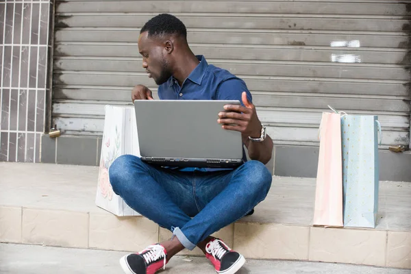 Młody Człowiek Siedzi Podłodze Laptopem Oglądając Torbie Zakupy — Zdjęcie stockowe