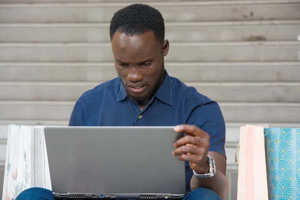 Młody Człowiek Siedzi Podłodze Torbami Zakupy Oglądając Laptop Patrząc Niezadowolony — Zdjęcie stockowe