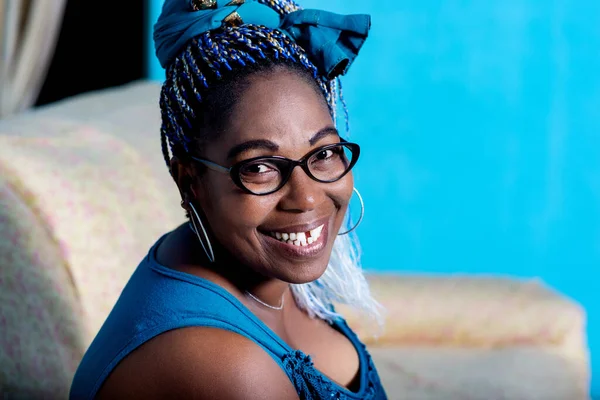 beautiful african adult woman dressed in blue dress with colorful braided hair and black eyeglasses, is sitting in the armchair of the house while being smiling