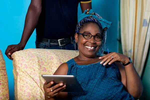 beautiful african adult woman dressed in blue dress with beautiful colorful braided hair sits in the armchair, holding in hand a digital tablet while being smiling