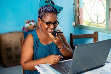 Güzel Afrikalı kadın bilgisayarla oturmuş cep telefonundan sesli mesaj yolluyor.