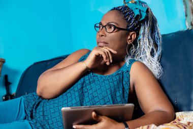 Gözlüklü güzel olgun bir kadın elinde tabletle kanepede uzanmış düşünüyor..