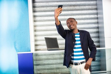 Yakışıklı Afrikalı yetişkin bir adam güneşli bir yaz gününde şehir sokağında beklerken cep telefonuyla selfie çekiyor. Açık havada akıllı telefondan online video görüşmesi yapan gülümseyen adam.