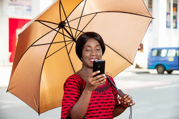 beautiful african adult businesswoman standing, holding an umbrella in hand, taking a self portrait photo in the street or chatting on video on her mobile phone smiling