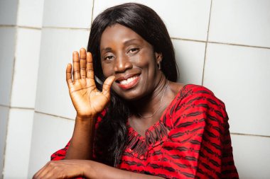 Geleneksel elbiseli güzel Afrikalı kadın bir duvarın yanında oturmuş kameraya gülümsüyor ve el hareketi yapıyor..