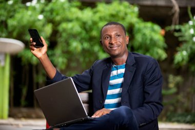 Genç adam parkta oturuyor, dizüstü bilgisayarla çalışıyor ve telefonunun ekranını gösteriyor.