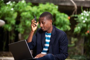 Genç adam parkta oturuyor ve yansıtıcı olarak dizüstü bilgisayar üzerinde çalışıyor.