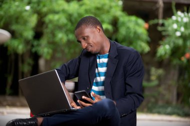 Yakışıklı iş adamı parkta oturmuş, elinde dizüstü bilgisayarla gülümsüyor.