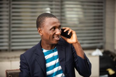 Mutlu ve yakışıklı Afrikalı yetişkin bir adam dışarıda bir bankta oturuyor ve gülümserken cep telefonunu arıyor.