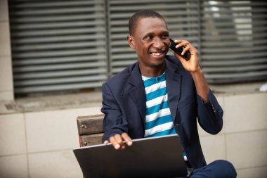 Yakışıklı, genç ve mutlu bir iş adamının bir bankta dizüstü bilgisayarla oturup cep telefonuyla konuşması..