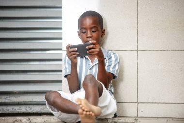 Küçük şirin çocuk dışarıda cep telefonuyla oyun oynuyor, gülümsüyor.