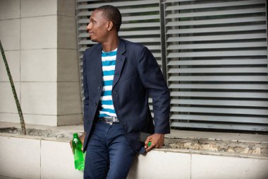 Şehir binalarının arka planında ceketli genç bir işadamının ciddi portresi ve şişe içinde su içmesi.