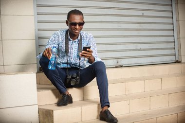 Siyah gözlüklü genç turist cep telefonuna bakıp gülümsüyor..