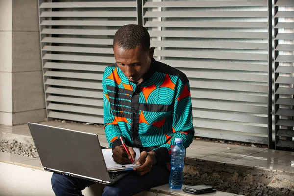 Счастливый Молодой Африканский Мужчина Сидящий Улице Записывающий Блокнот Помощью Ноутбука — стоковое фото