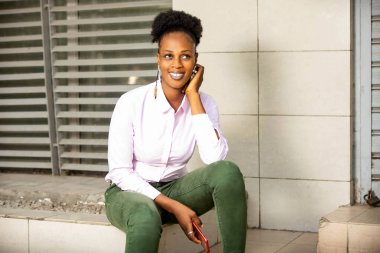 Beyaz gömlek ve yeşil kot pantolon giyen güzel genç bir kadın merdivenlerde oturuyor ve gülümserken cep telefonunu tutuyor.