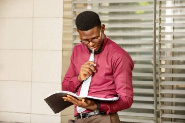 Genç Afrikalı bir adam ayakta, bir kütük tutuyor ve ileriye bakıyor ağzında bir stelo tutarak; güzel bir bordo kırmızı kimya ve bezel giyiyor