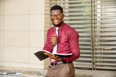 Genç Afrikalı bir adam ayakta, elinde bir kütük tutarken gülümseyen bir stelo gösteriyordu; güzel bir bordo kırmızı kimya ve bezel giyiyordu.