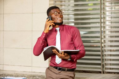 Genç Afrikalı elinde bir kayıt tutarak gülümsüyor ve üzerinde güzel kırmızı bordo bir kimya ve gözlük ile bir telefon görüşmesi yapıyor.