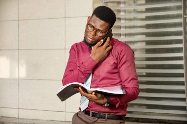 Genç iş adamı dışarıyı ararken cep telefonunu kullanıyor. Kırmızı bordo giyen ve elinde defter tutan genç bir adam.