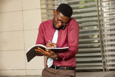 Genç, meşgul bir Afrikalı adam aynı anda iki şey yapıyor telefonla konuşuyor ve defterine not alıyor. İşini planlamak için telefonu omzundan tutan endişeli bir adam. Kravatlı kırmızı bir kimya giyiyordu.