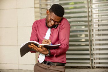 Genç, meşgul bir Afrikalı adam aynı anda iki şey yapıyor telefonla konuşuyor ve defterine not alıyor. İşini planlamak için telefonu omzundan tutan endişeli bir adam. Kravatlı kırmızı bir kimya giyiyordu.