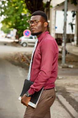 Genç gülümseyen Afrikalı öğrenci akademik arkaplanda elinde bir not defteri tutuyor. Açık havada gülümseyen kendinden emin genç bir öğrenci. Kampüsteki öğrenci kolunu uzatıyor. Kırmızı bir kimya ve kravat taşıyordu..