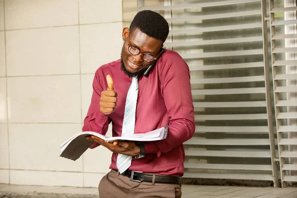 Meşgul genç Afrikalı bir adam aynı anda iki şey yapıyor, telefonda konuşuyor ve defterini tutarken el kol hareketi yapıyor. İşini planlamak için telefonu omzundan tutan endişeli bir adam. Kravatlı kırmızı bir kimya giyiyordu.