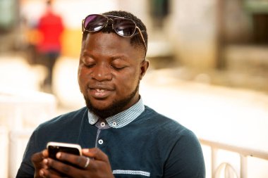 Genç Afrikalı bir adam elinde telefonuyla otururken alnına güneş gözlüğü takıyor.