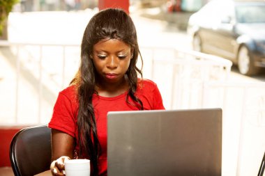 Güzel genç bir kadın verandasında oturmuş dizüstü bilgisayarını kullanıyor ve elinde bir bardak tutuyor..