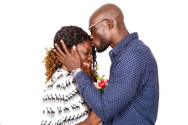 Gözlüklü genç adam beyaz arka planda duruyor kız arkadaşının alnından öpüyor elinde bir çiçekle gülüyor.