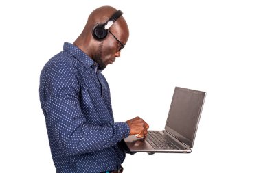 Beyaz arka planda duran yakışıklı bir işadamı kulaklık kullanarak müzik dinlerken dizüstü bilgisayarla çalışıyor.