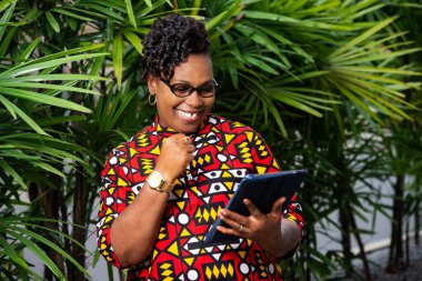 Geleneksel peştamallı, dijital tabletin yardımıyla ayakta duran mutlu ve güzel yetişkin bir kadın zafer jesti yapıyor.