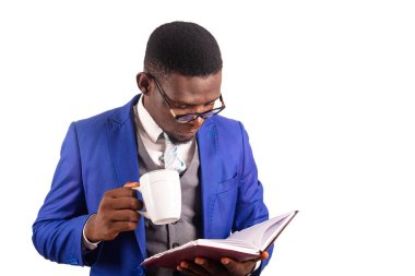 Ceketli genç işadamı beyaz arka planda tek başına duruyor ajandasını kontrol ediyor ve elinde bir fincan kahve tutuyor.