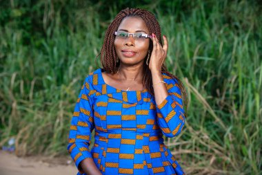 Geleneksel elbiseli, güzel, olgun bir kadın parkta durmuş gözlüklerine dokunuyor ve gülümseyen kameraya bakıyor..