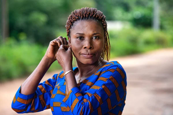 a beautiful African woman in traditional dress standing in the countryside adjusting her earring while smiling.