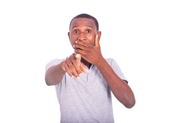 Beyaz arka planda duran tişörtlü genç bir adam eliyle ağzını kapatan bir şey gösteriyor..