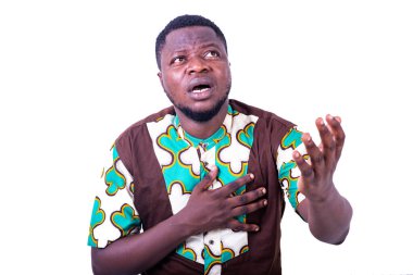 Peştamal gömlekli genç adam beyaz arka planda duruyor, elleri göğsünde yukarı bakıyor ve el hareketi yapıyor..