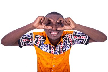 Beyaz arka planda duran kolsuz gömlekli yakışıklı genç bir işadamı dürbün gibi duruyor ve parmaklarının arasından gülümsüyor.