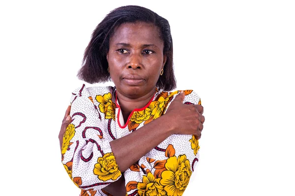 Beyaz bir arka planda duran ve kollarını kavuşturarak kameraya bakan, çiçek takmış olgun bir kadın.