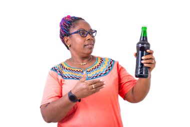Beyaz arka planda duran gözlüklü Afrikalı bir kadın konuşurken bir şişe biraya bakıyor..
